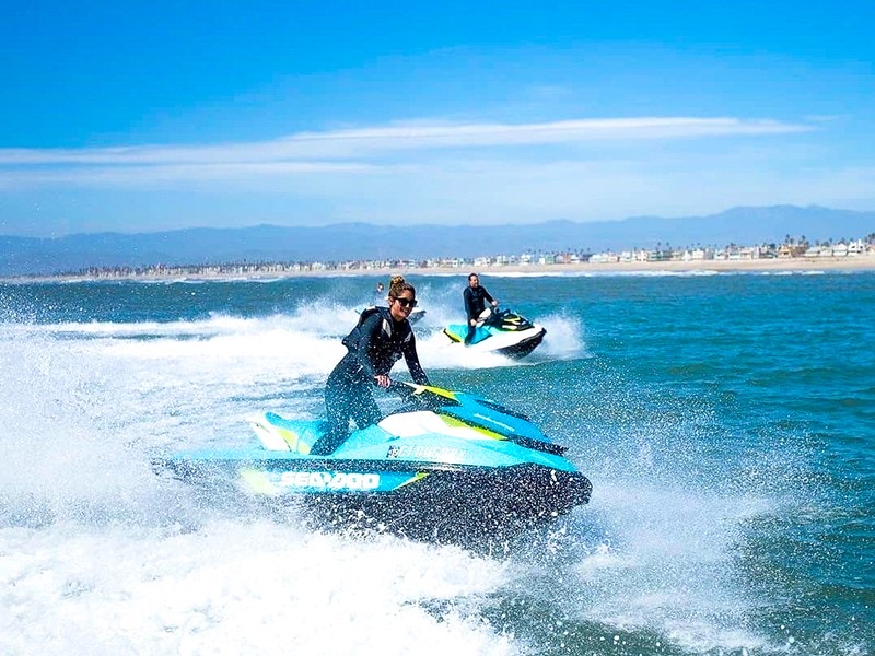 Two riders on personal watercraft kicking up spray near a beach